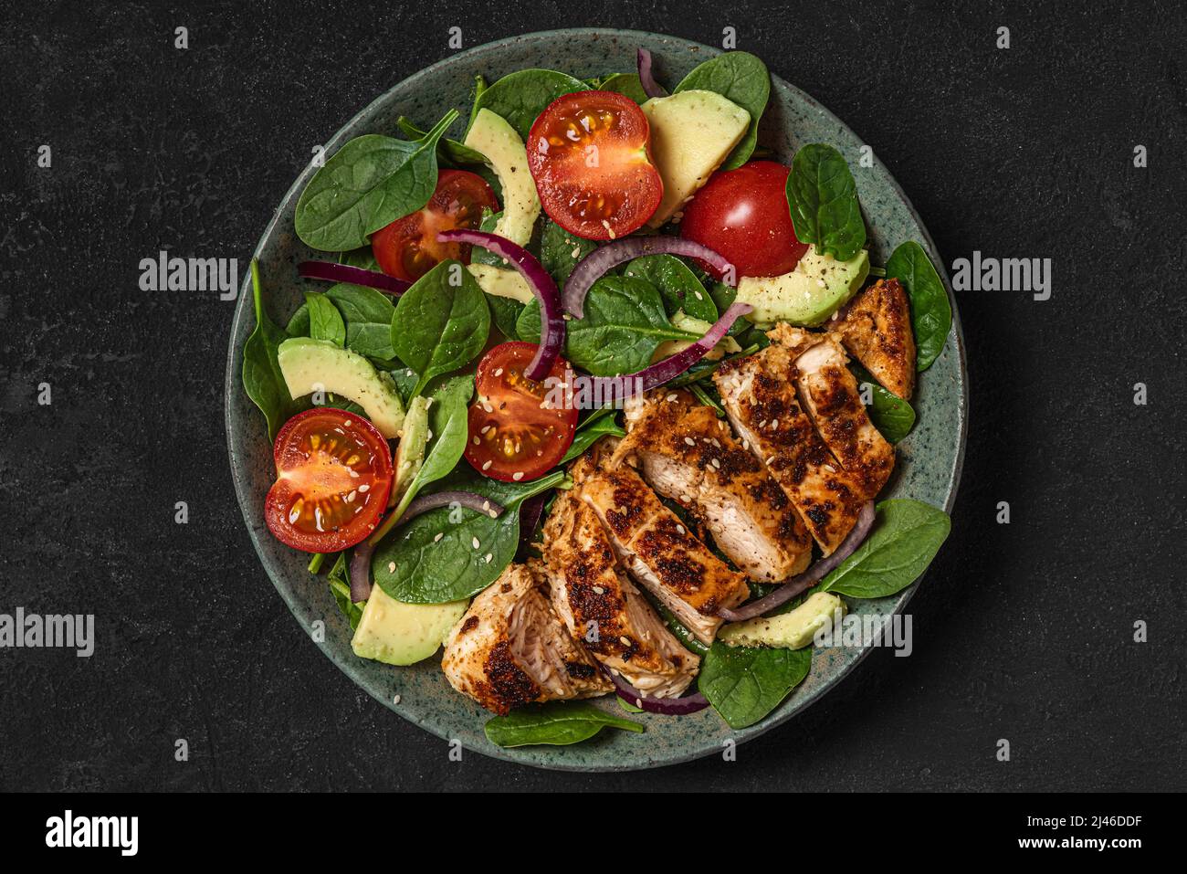 Grilled chicken breast and spinach salad with avocado, tomatoes and sesame seeds in a plate on dark background. top view Stock Photo