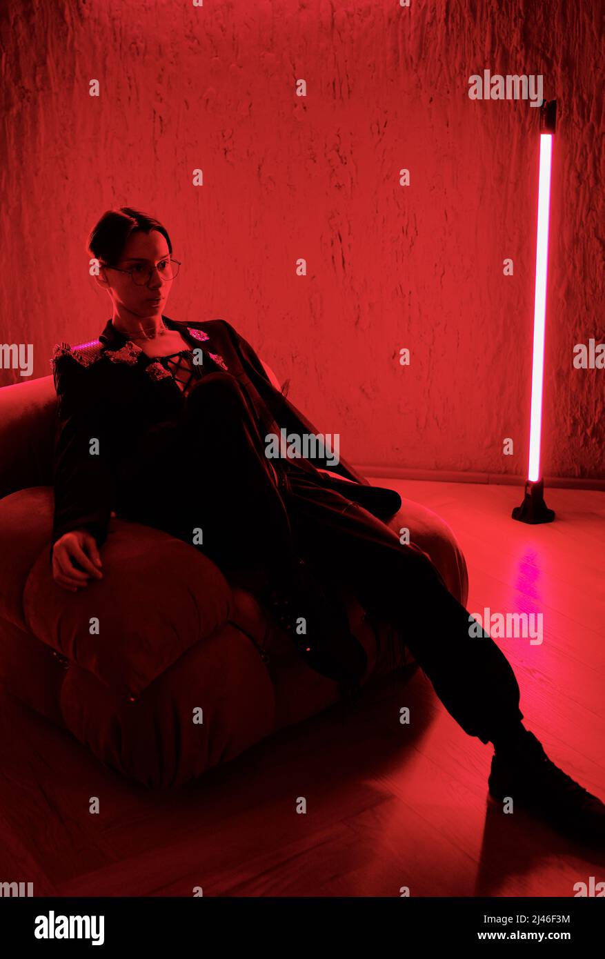 Young restful posh man or teenage guy in smart suit relaxing in armchair in night club in spacious stylish room lit with red light Stock Photo