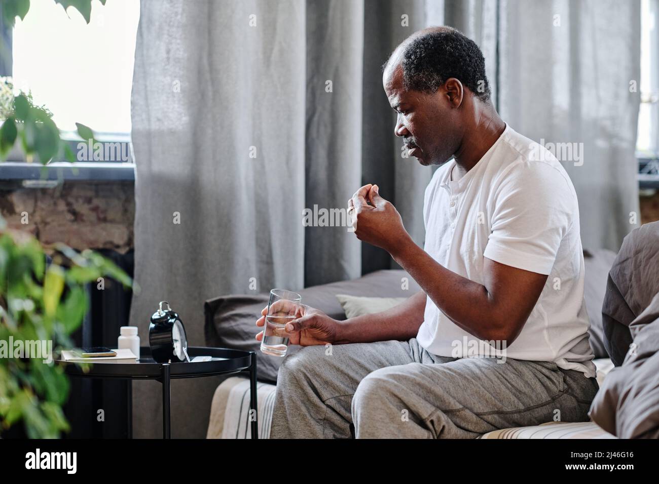 Senior black man with glass of water taking medicaments prescribed by his general practitioner while sitting on bed after or before sleep Stock Photo