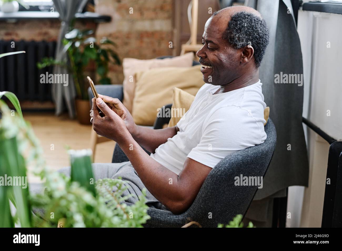 Contemporary senior black man in home wear communicating in video chat while sitting in comfortable armchair in living room Stock Photo