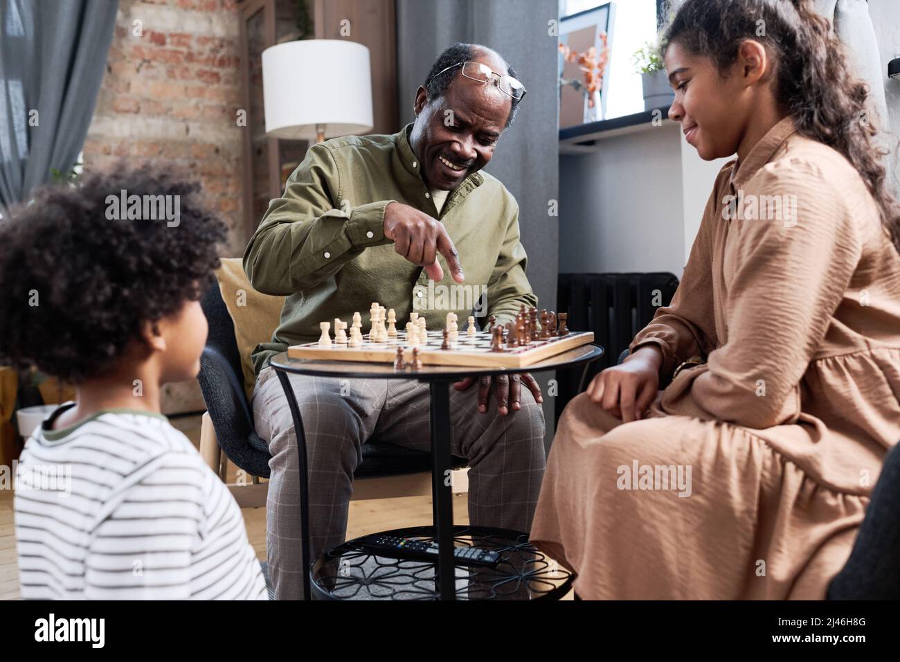 Happy retired black man pointing at pawn on chess board while explaining his grandchildren rules of leisure game Stock Photo