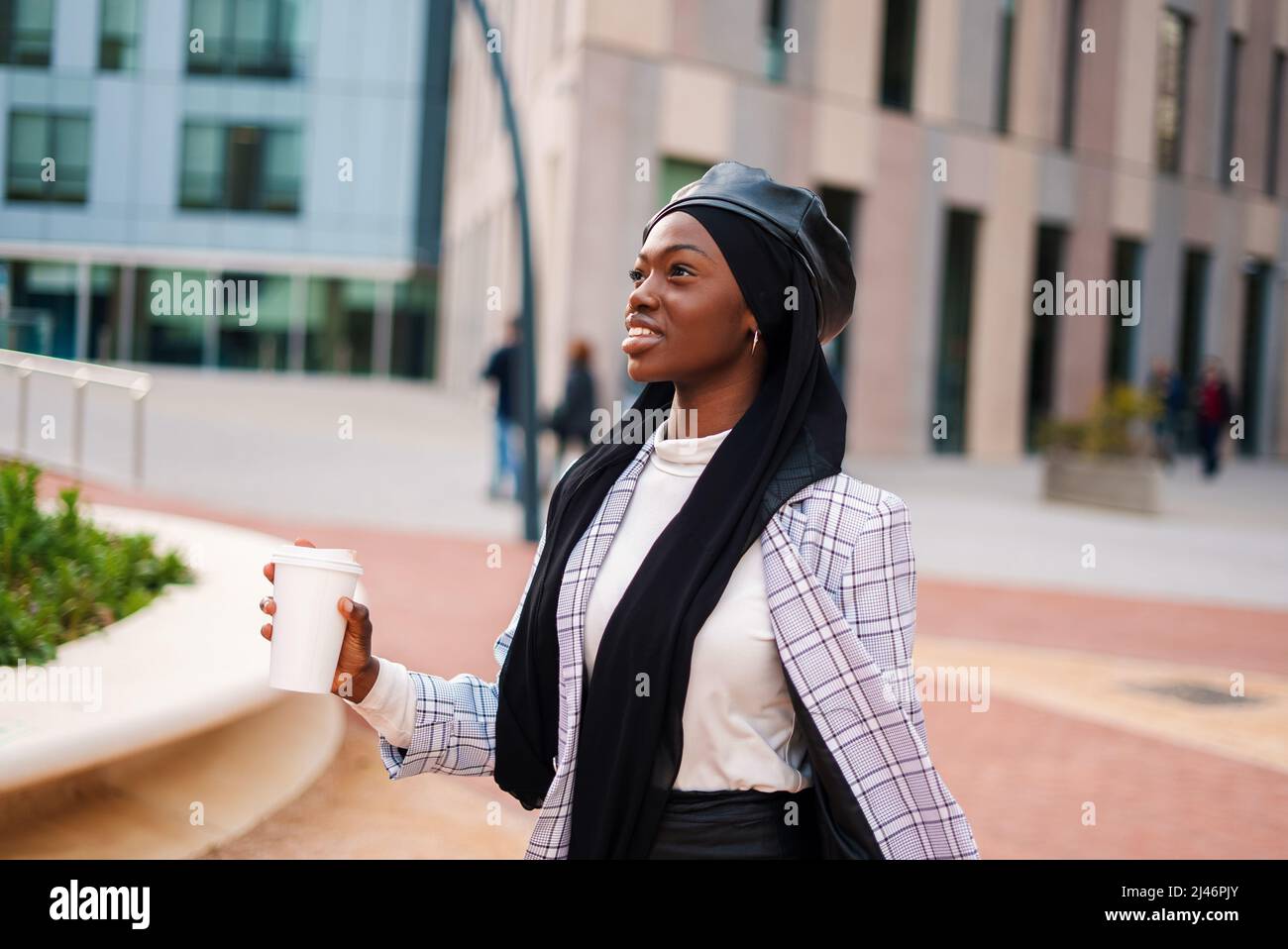 Happy African American woman in smart casual clothing and Muslim headscarf carrying takeaway coffee in public city park Stock Photo