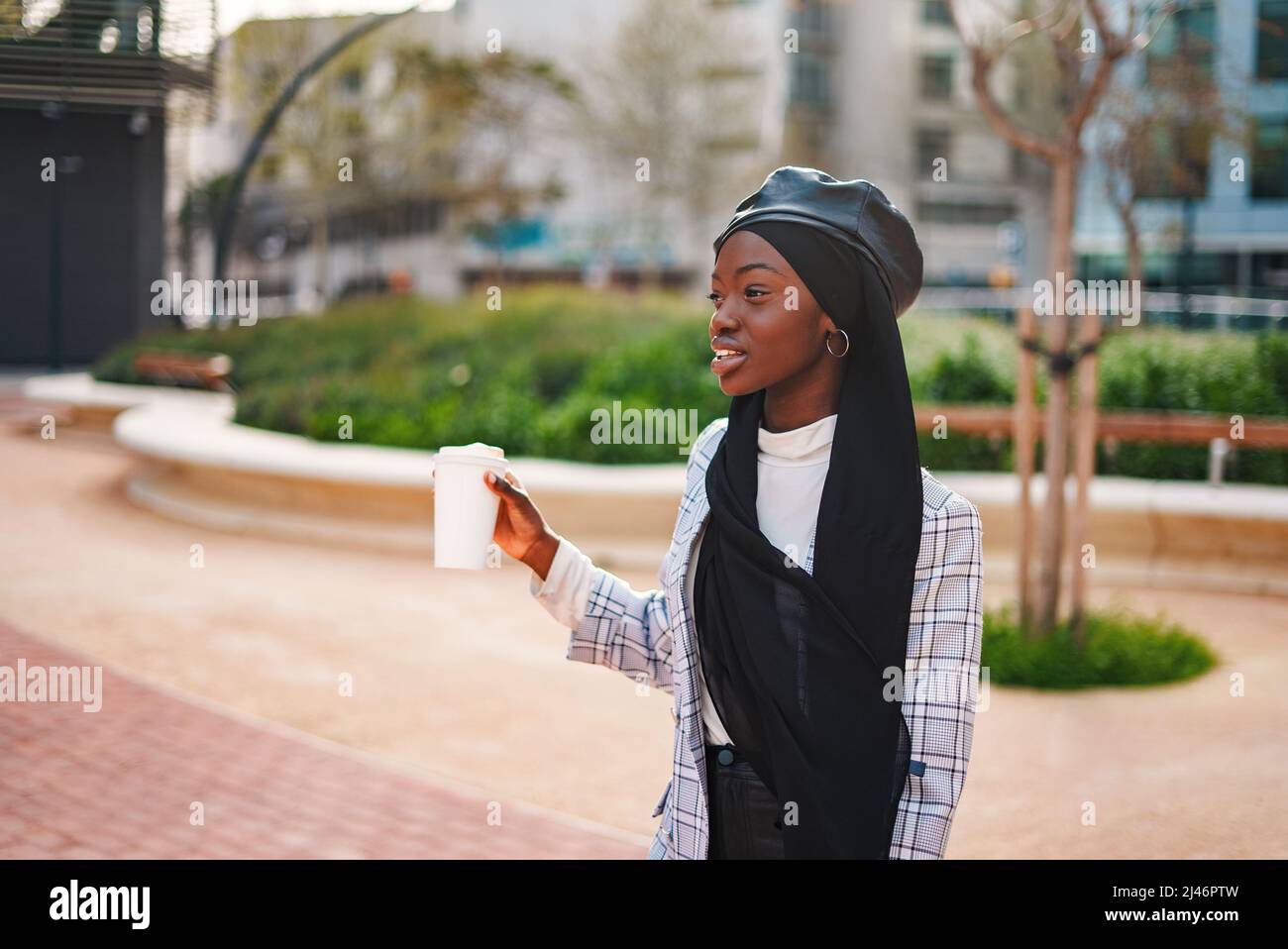 Happy African American woman in smart casual clothing and Muslim headscarf carrying takeaway coffee in public city park Stock Photo