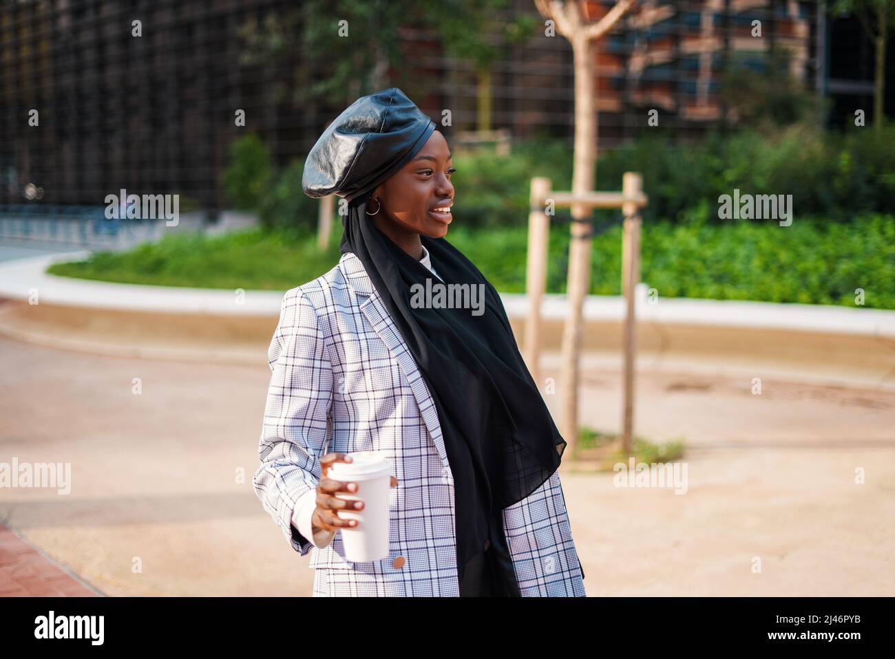 Positive African American female dressed in smart casual clothes and traditional headscarf standing with disposable cup of hot takeaway beverage and looking away Stock Photo