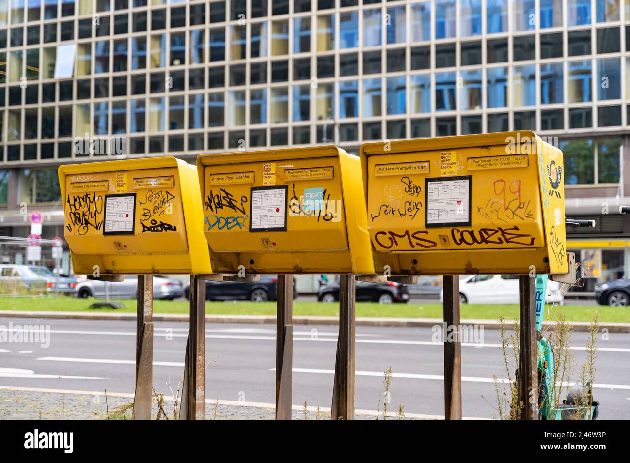 Three yellow postboxes from the Deutsche Post next to a big street in the city. Mailboxes to send letters and cards. Graffiti tags and sticker. Stock Photo