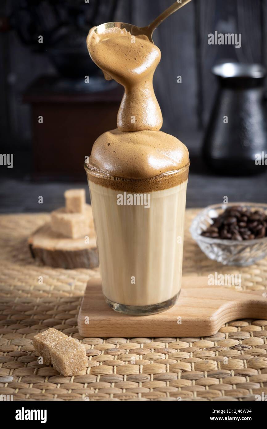 Iced Dalgona Coffee, a trendy fluffy creamy whipped coffee, on dark wooden background with coffee grinder, sugar and coffee beans, with foam spoon Stock Photo