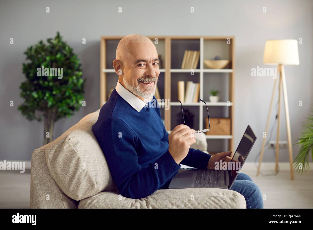 Happy, smiling senior man sitting on couch at home and using his laptop computer Stock Photo