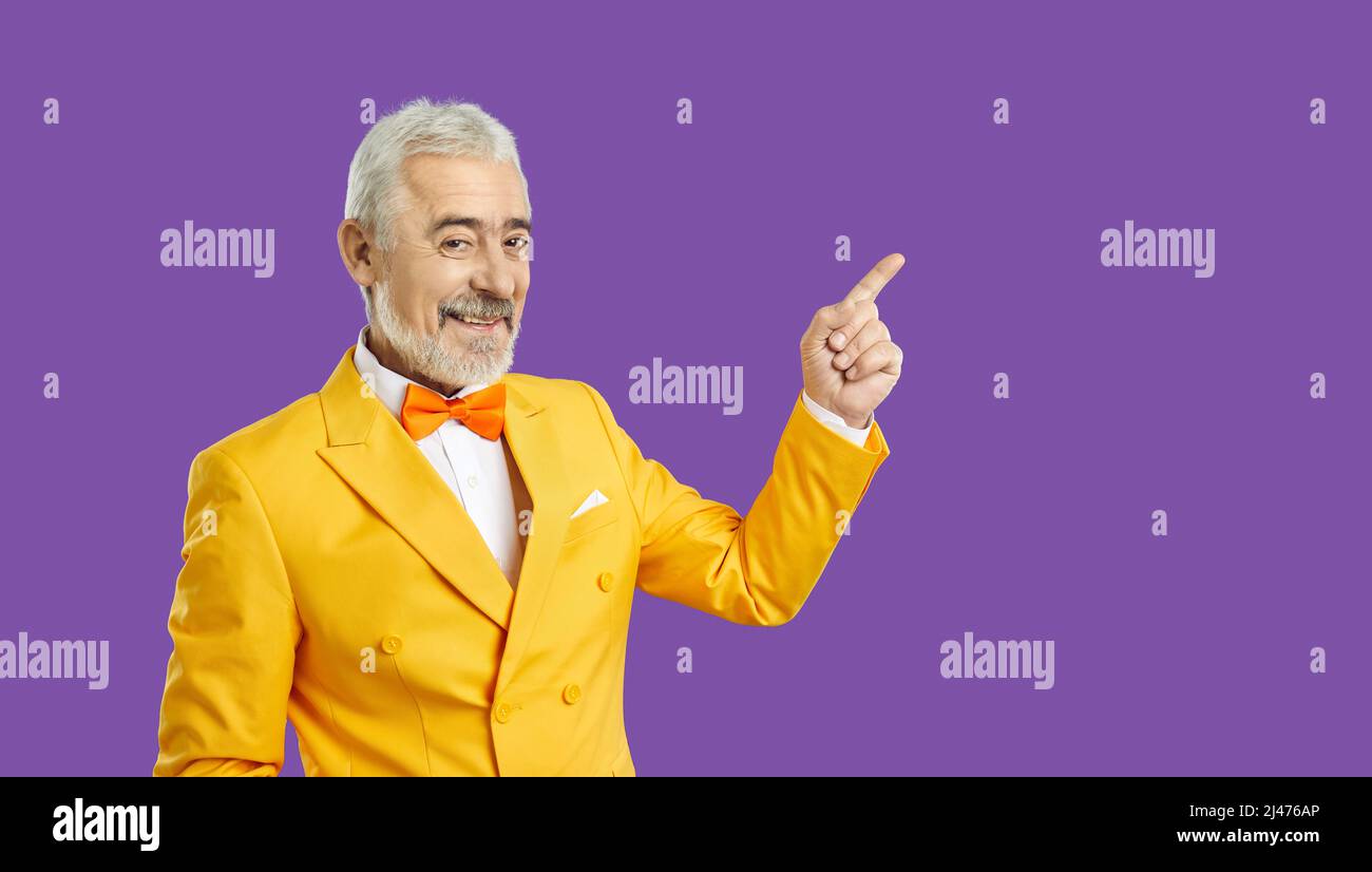Happy senior man in yellow suit on purple background smiles and points his finger at copy space Stock Photo