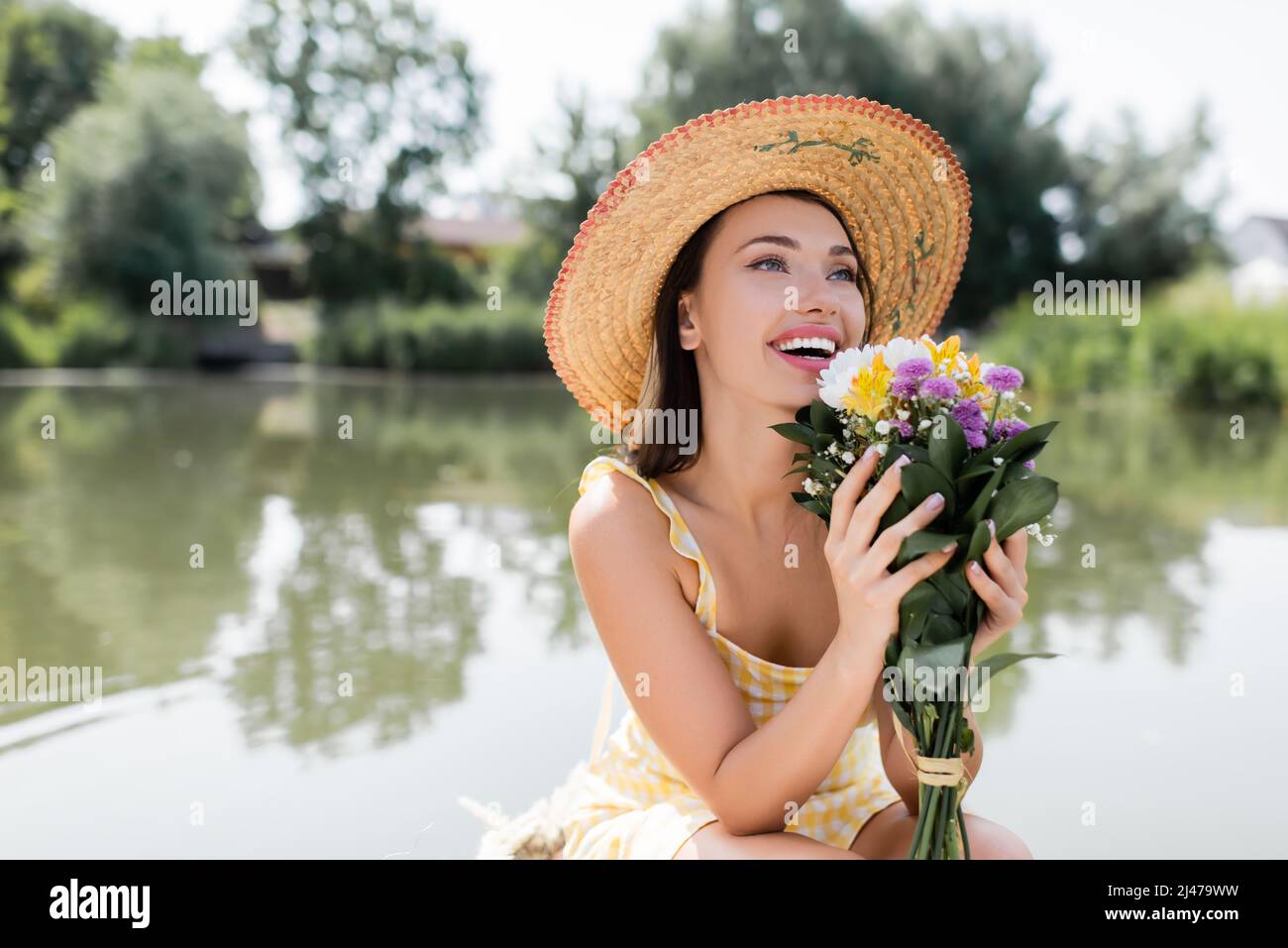 joyful young woman in straw hat and dress holding flowers near lake ...