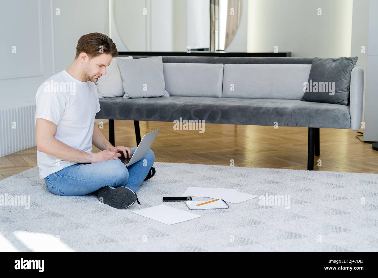 full length of man sitting on floor in living room and typing on laptop ...