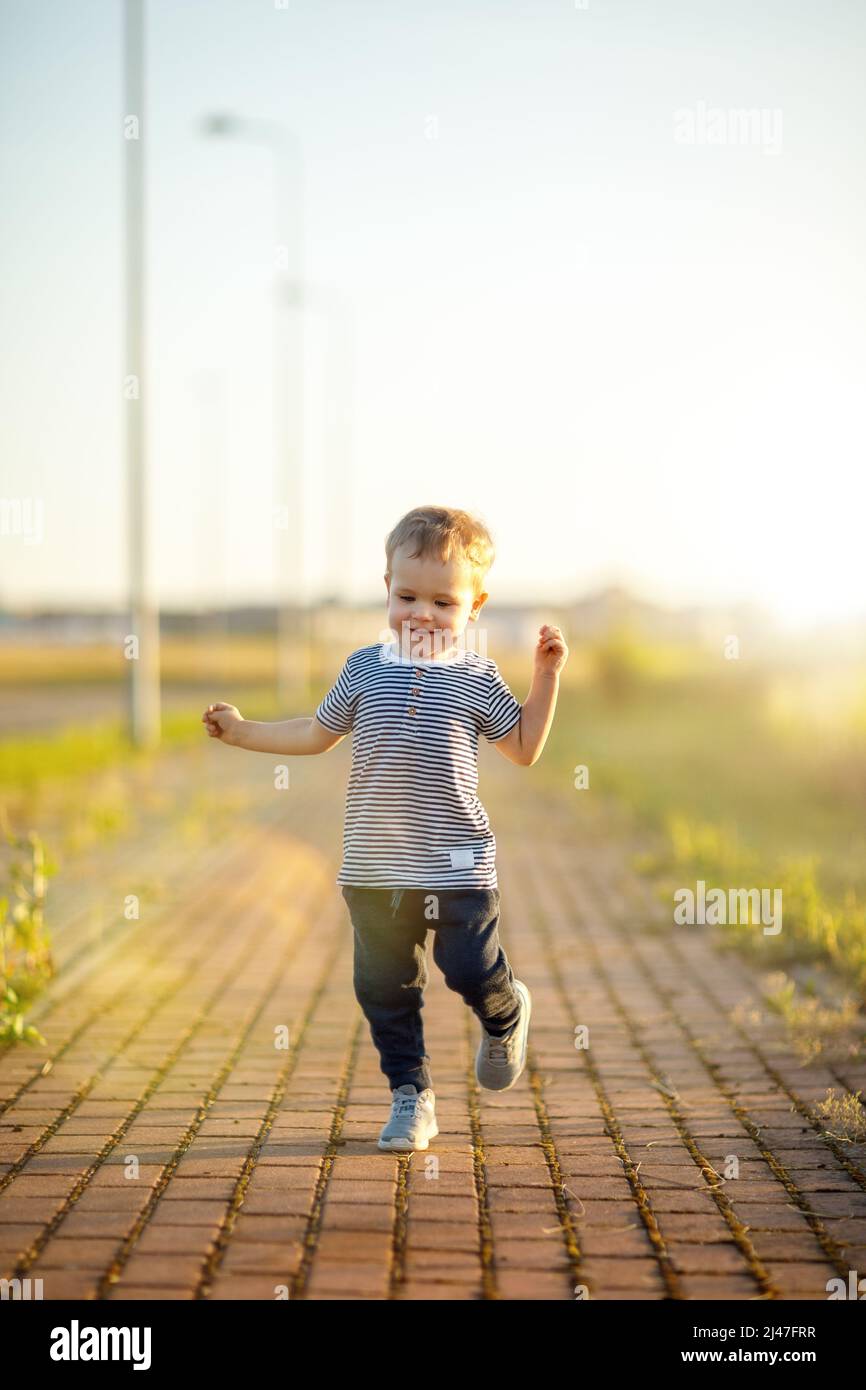 Cheerful little boy with striped shirt runs down the sidewalk. Sunny summer evening, backlighting. Stock Photo