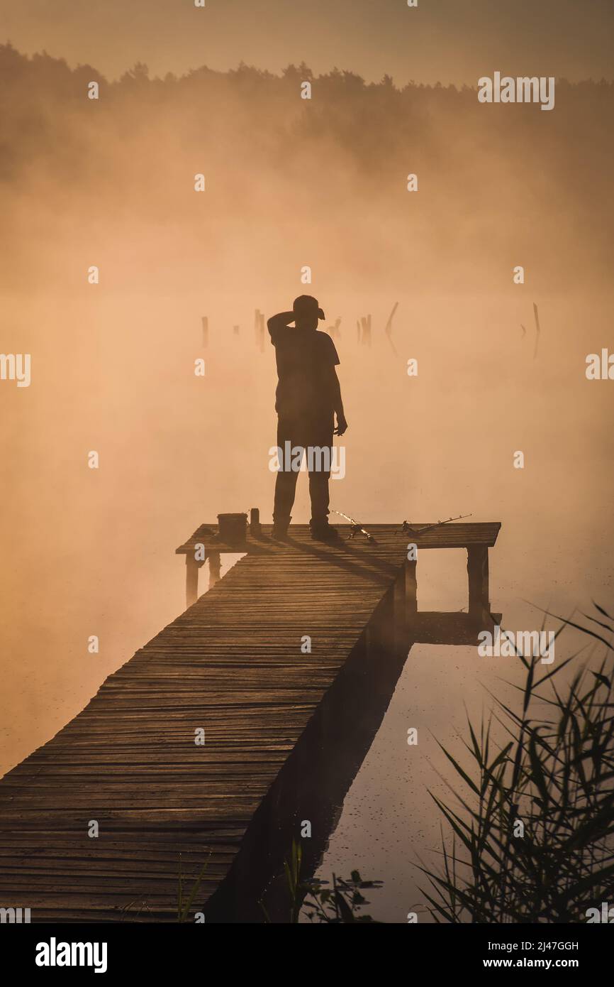 Beautiful morning scene by the lake. Silhouette of a man fishing on a wooden pier in the light of the rising sun. Stock Photo