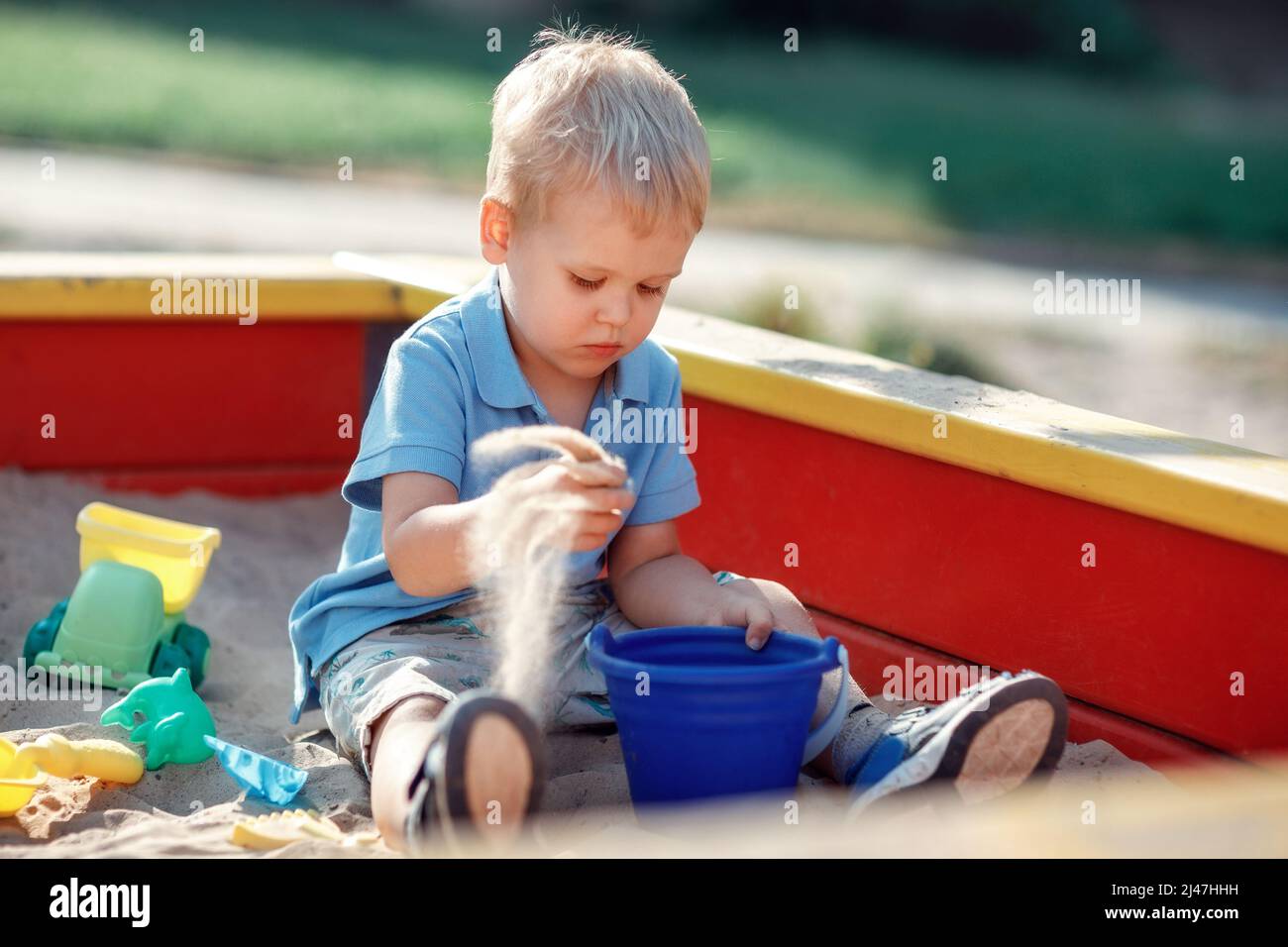 Toddler boy is playing in a red and yellow edge sandbox with sand toys. Focused baby, wearing blue shirt is trying to put sand in a blue bucket. Child Stock Photo