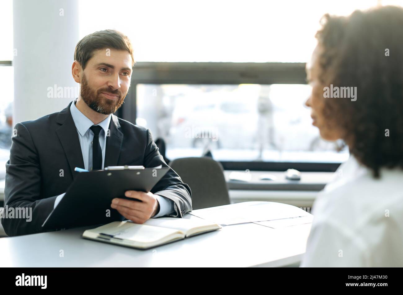 Interview. Young adult caucasian male, hiring manager, sits at a desk in a modern office, conducts an interview with an African American girl, looks through a resume, talks about working conditions Stock Photo