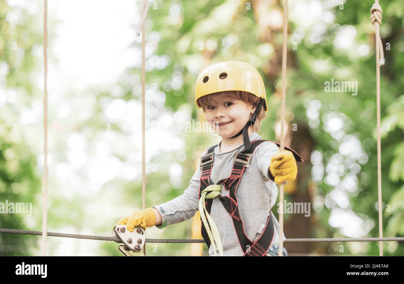 Happy child boy calling while climbing high tree and ropes. Portrait of a beautiful kid on a rope park among trees. Small boy enjoy childhood years. Stock Photo