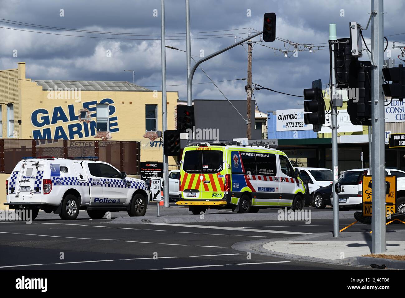 Ambulance and police vehicle stopped at an intersection in the suburbs of Melbourne, with shops and dark clouds in the background Stock Photo