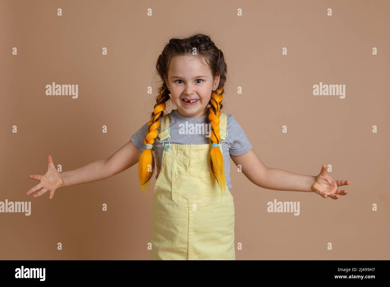 Indulging young girl with yellow kanekalon pigtails, scaring someone with weird grimace and hands wearing yellow jumpsuit and gray t-shirt on beige Stock Photo