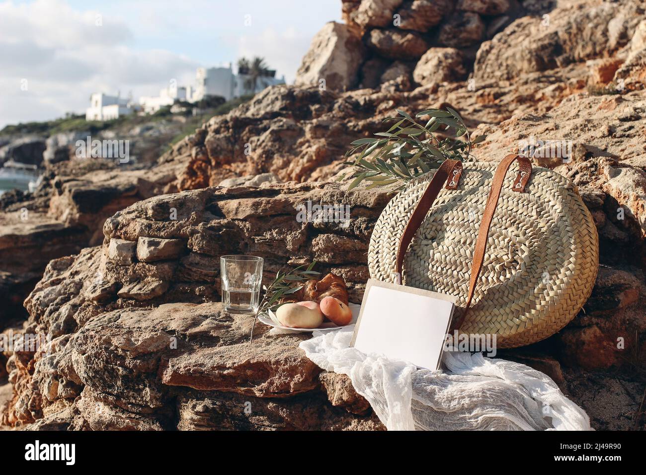 Summer vacation. Picnic breakfast on beach. Straw basket, olive tree branches. Blank mockup card. Glass of water, peach fruit. Blurred background with Stock Photo