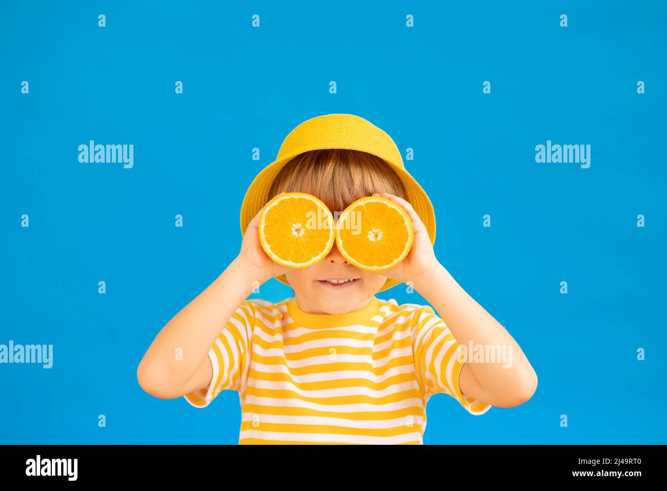 Happy child holding slices of orange fruit like sunglasses. Kid wearing striped yellow t-shirt against blue paper background. Healthy eating and summe Stock Photo