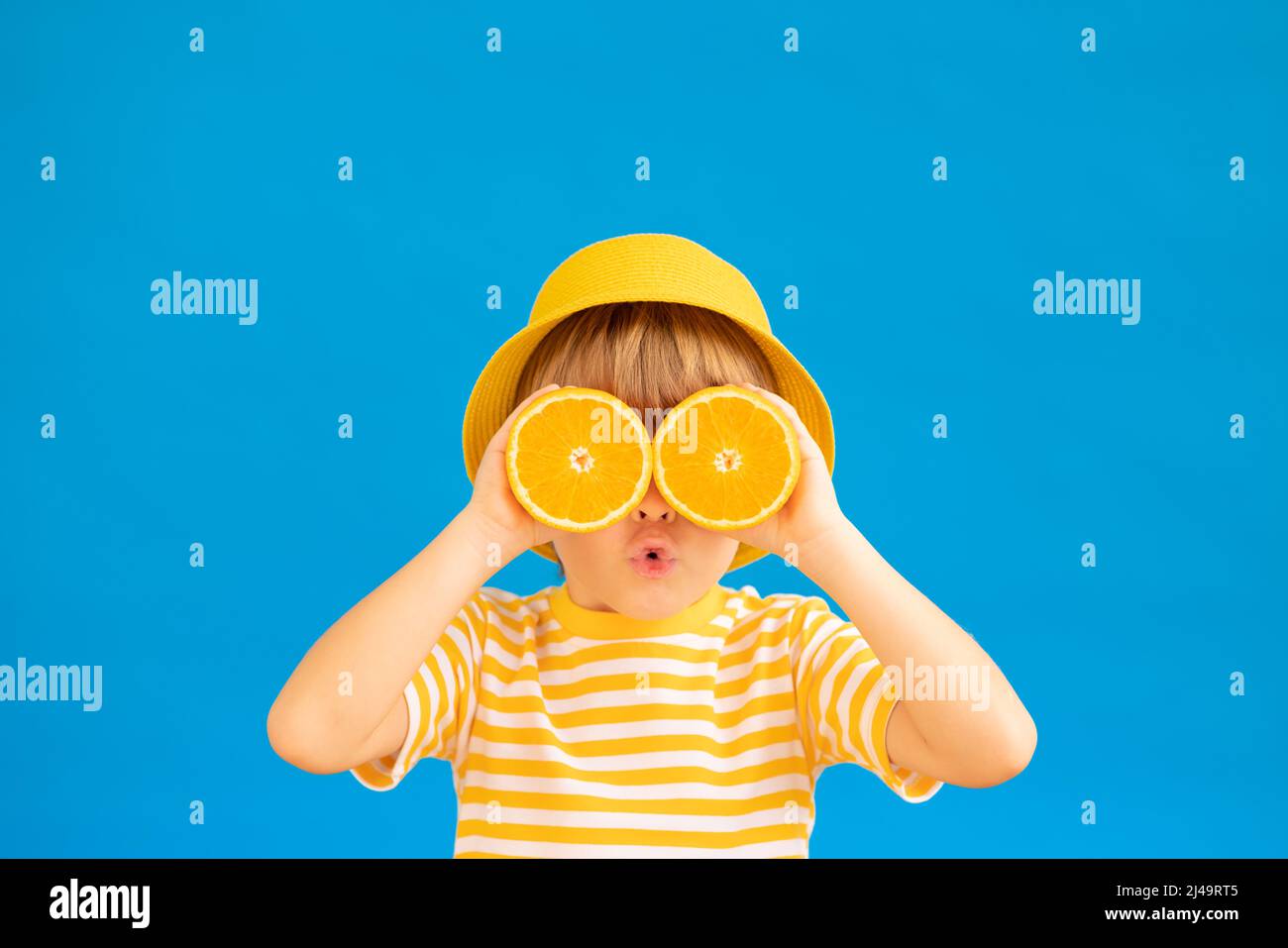 Surprized child holding slices of orange fruit like sunglasses. Kid wearing striped yellow t-shirt against blue paper background. Healthy eating and s Stock Photo