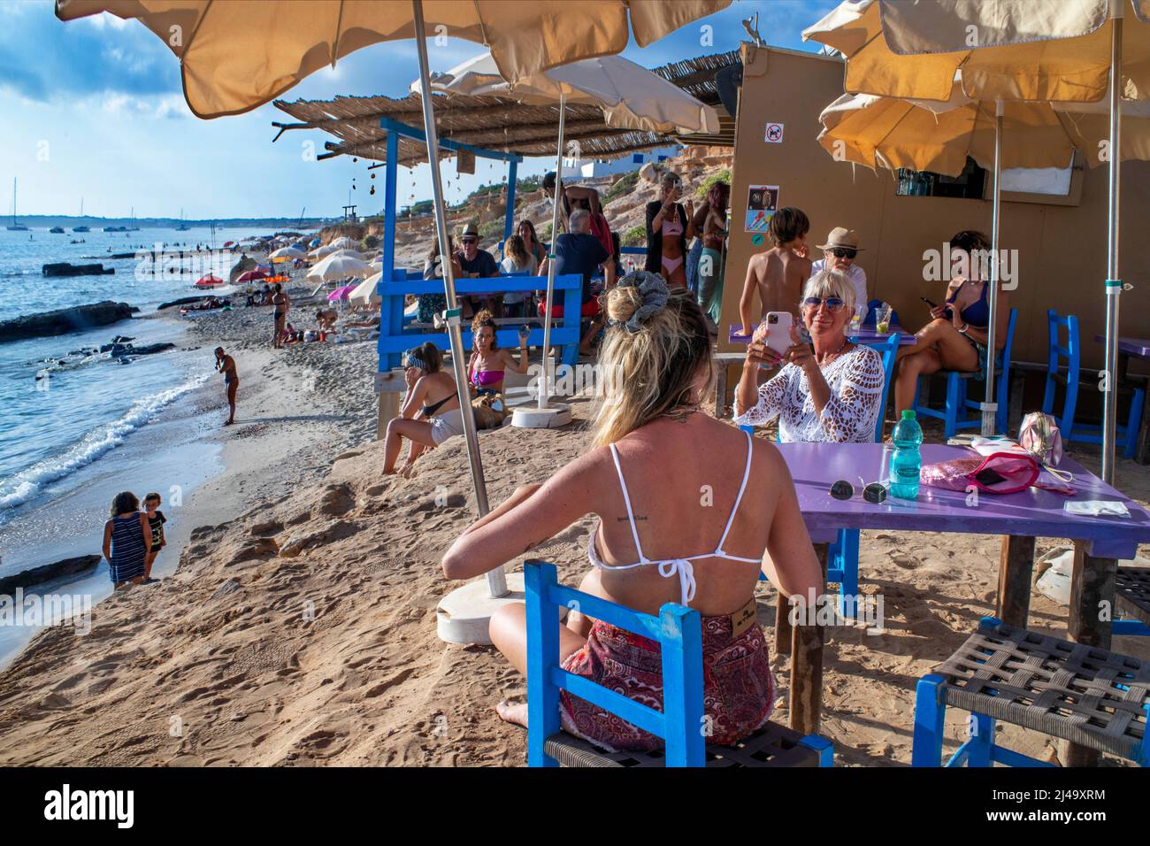 Chiringuito Bartolo bar restaurant in Playa Mitjorn beach in es Caló d´es mort cala beach, summer visitors lying on a cove lined with red cliffs and r Stock Photo