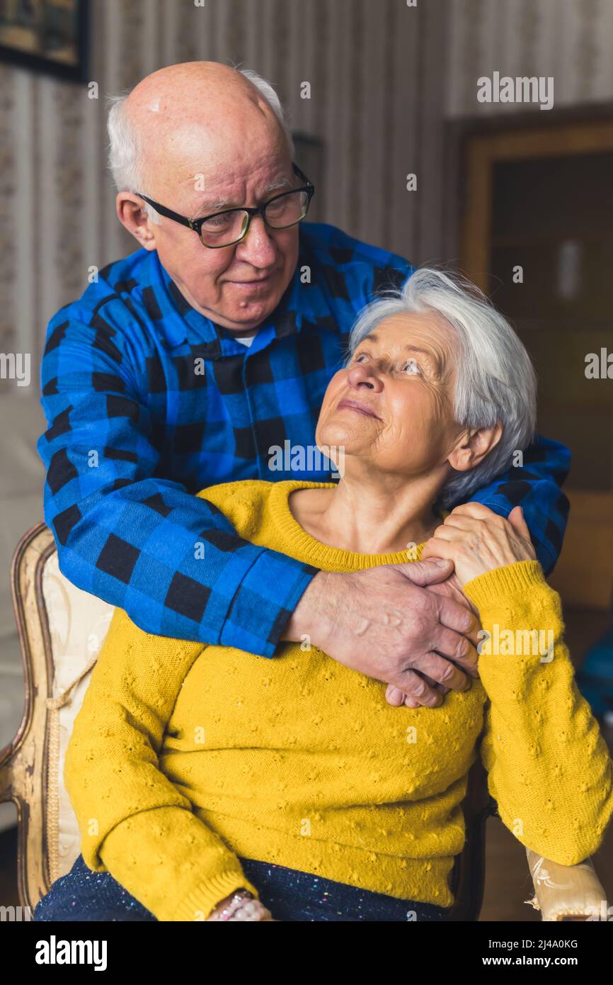 Vertical shot of a hugging elderly caucasian couple. Senior gray-haired woman sitting on an old-fashioned chair being embraced by her bald charming husband in a flannel shirt. High quality photo Stock Photo