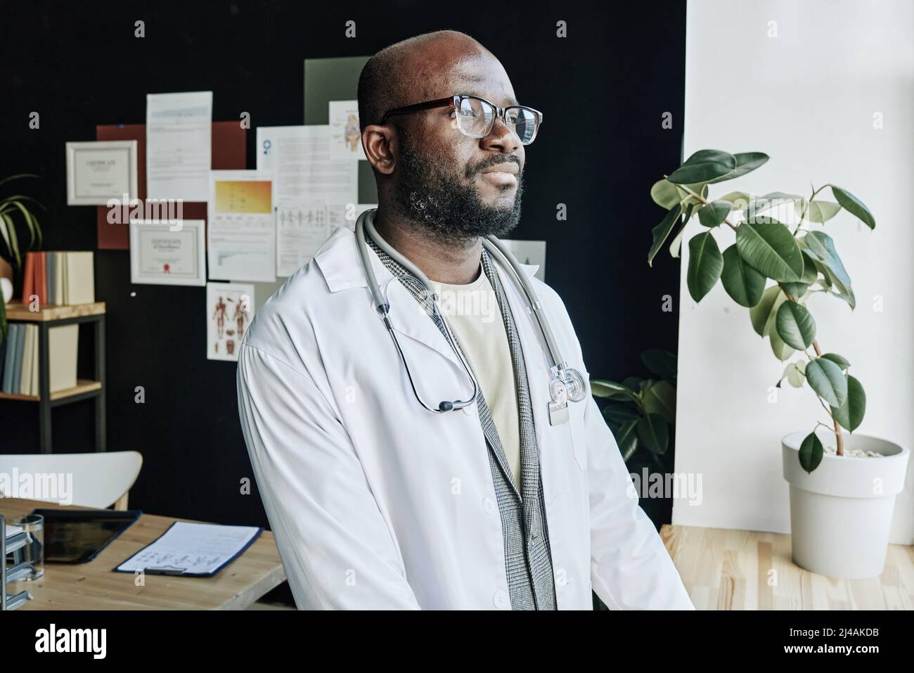 African male therapist in white coat and eyeglasses looking at window with smile on his face during work at office Stock Photo