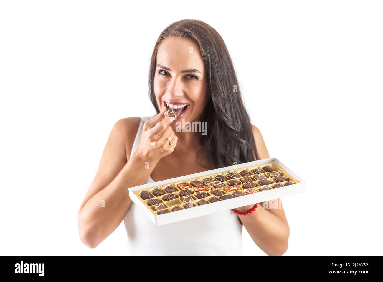 Happy dark-haired woman is taking a bite from a sweet candy while holding a full candy box. Isolated white background. Stock Photo