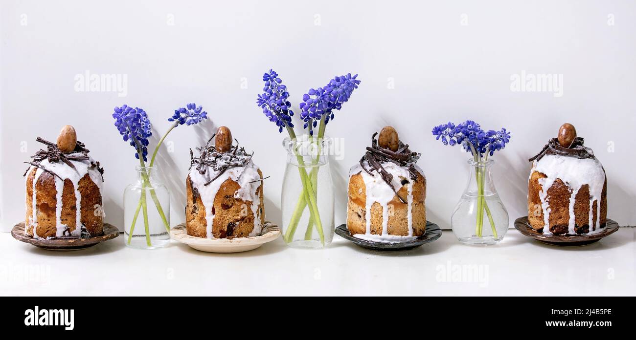 Homemade traditionla small Easter kulich cakes with chocolate nests and eggs on plates in row decorated with muscari flowers over white marble table. Stock Photo