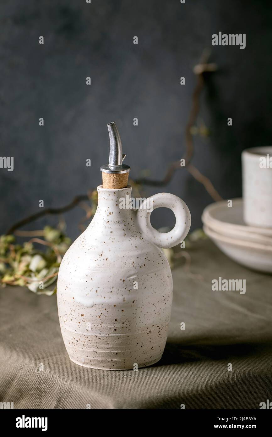 White speckled craft ceramic olive oil or vinegar bottle dispenser standing on dark green linen tablecloth with stack of plates. Rustic style, coly sp Stock Photo