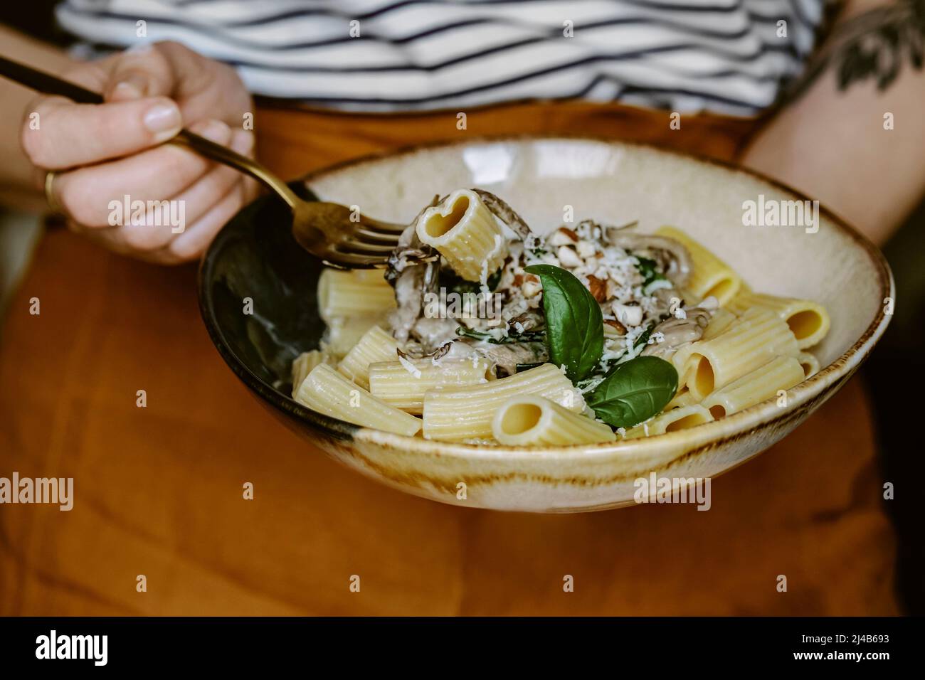anonymous woman with linen terracota apron, holding a Rigatoni pasta dish, with cream, mushroom, spinach, almond and grated parmesan, in handmade plat Stock Photo