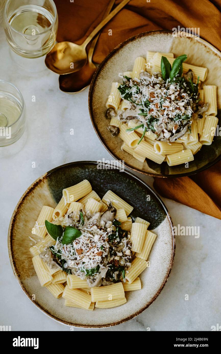 Rigatoni pasta dish, with cream, mushroom, spinach, almond and grated parmesan, in handmade plates, and glass of white wine, ocher linen tablecloth, g Stock Photo