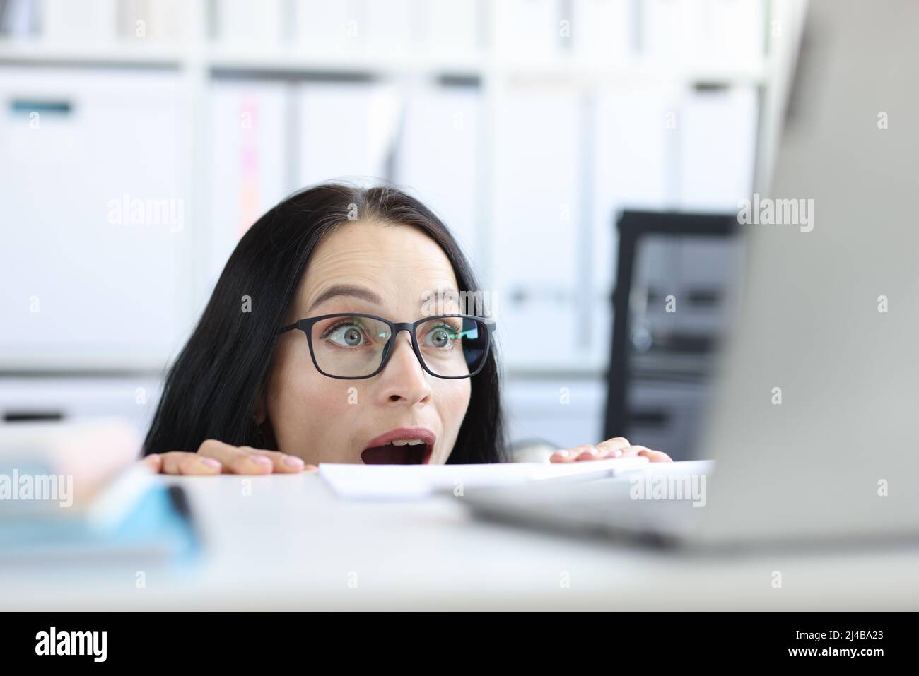 Young businesswoman peering over her desk in office in wide eyed horror or amazement Stock Photo