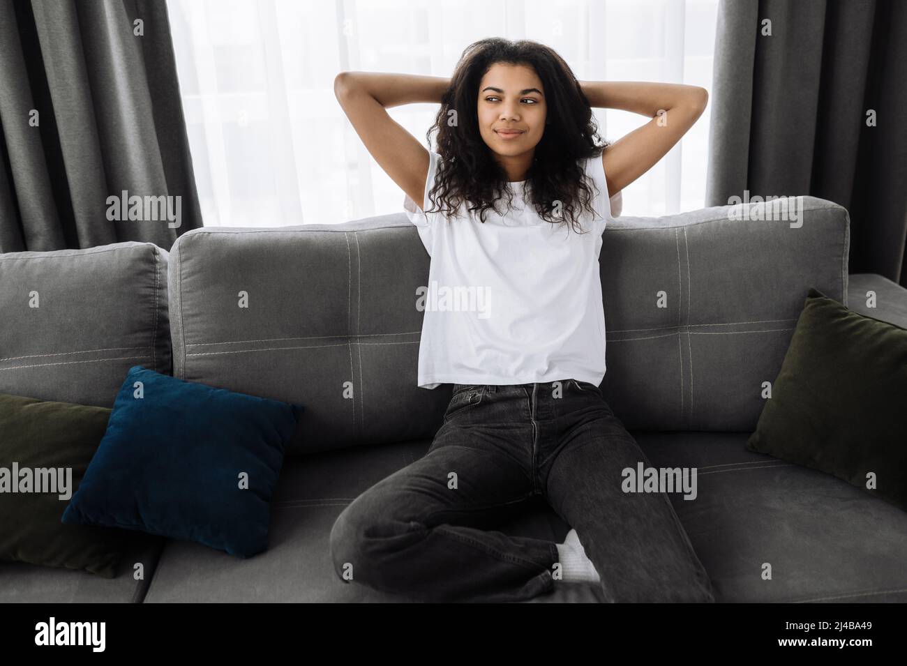 A beautiful young woman is relaxing on the couch in living room at home, smiling. African american girl takes a break from online distant work, or have lazy leisure. Happy attractive female dreaming Stock Photo