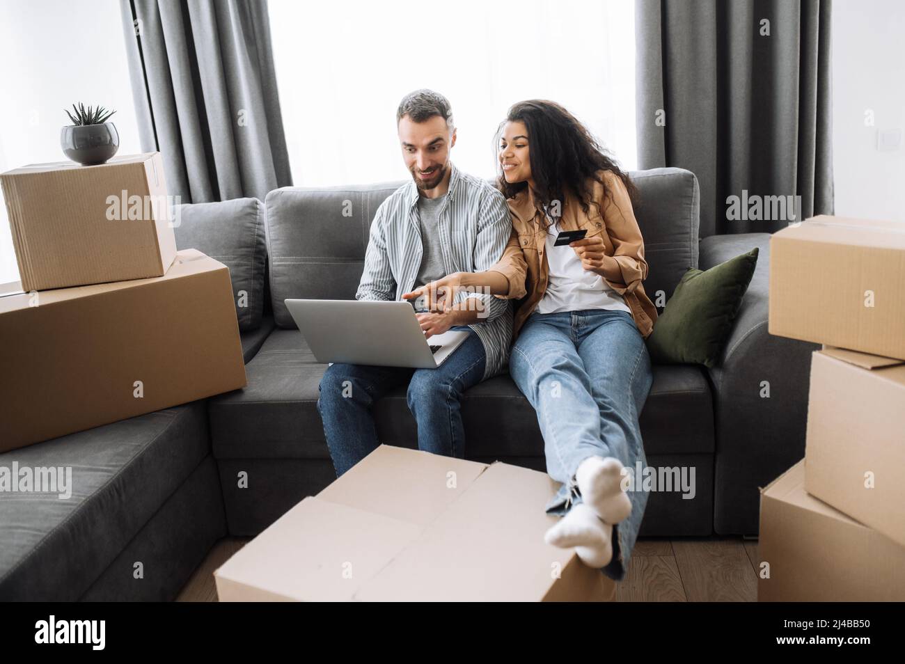 Happy couple beween a heap of boxes in living room sitting at the couch use a laptop and credit card for buying on-line, choose furniture on webstore, search design interior ideas fot their new home Stock Photo