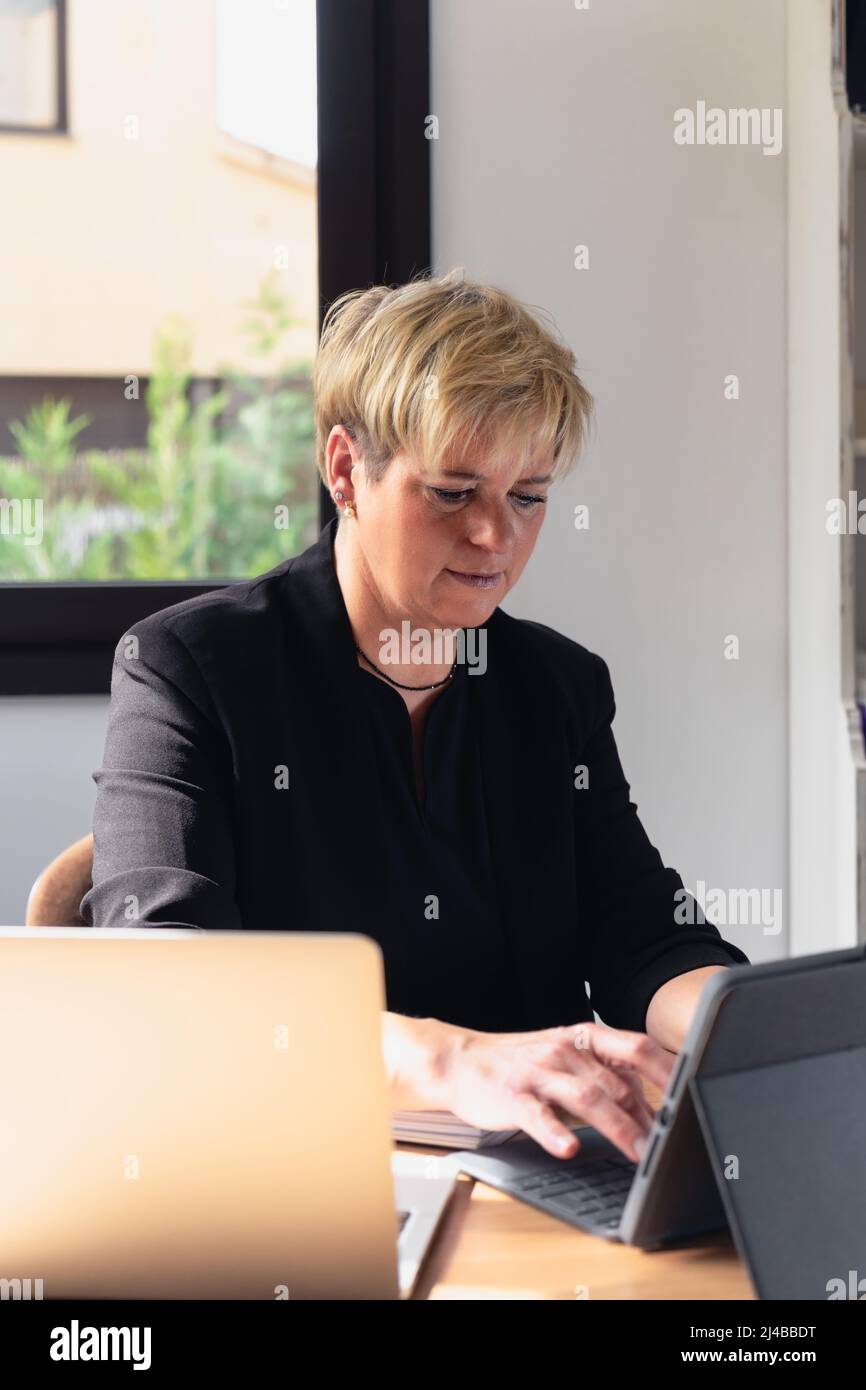 Mature business woman with blonde hair, working in her home office talking on the phone while consulting her laptop. Stock Photo