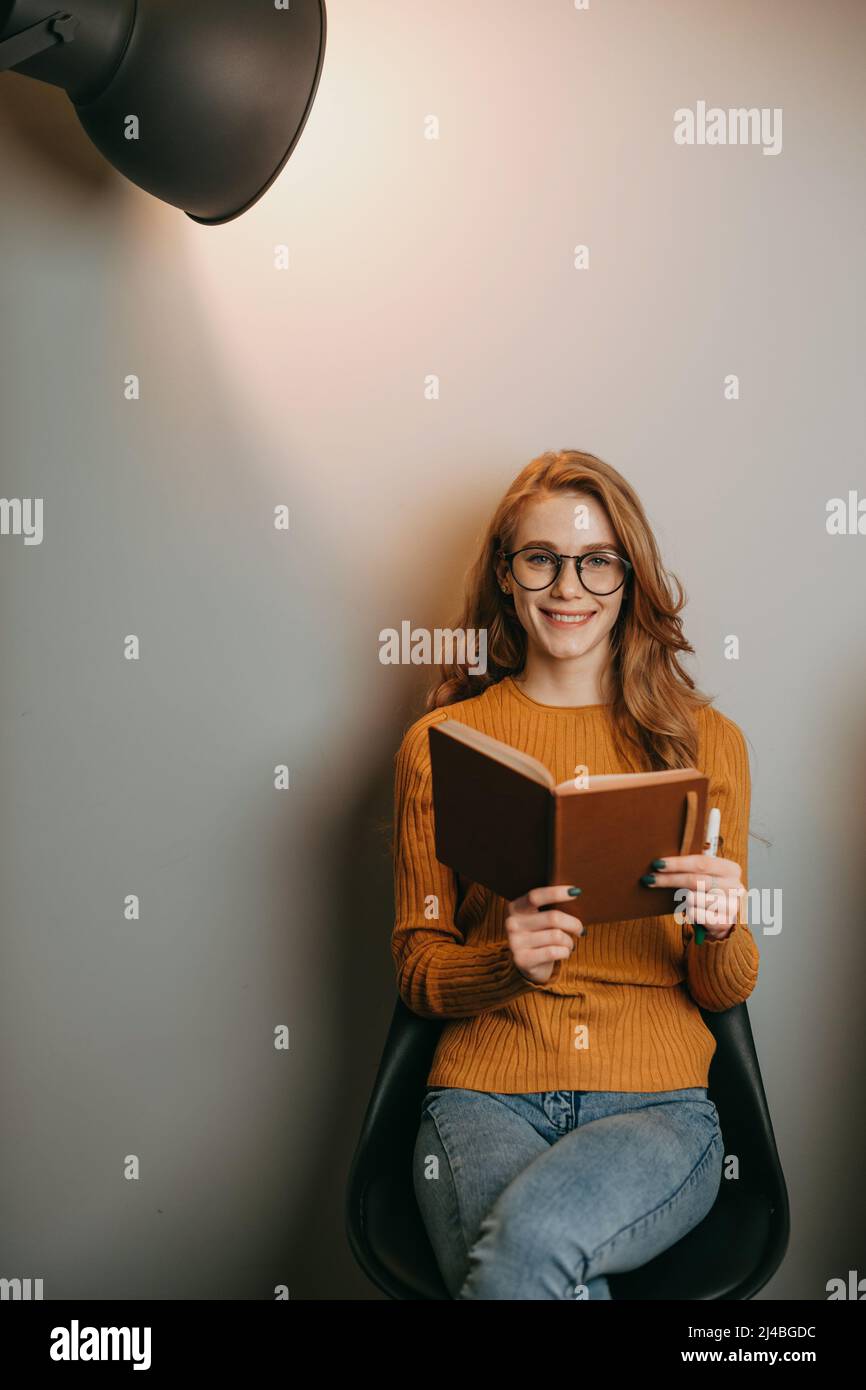 Red-headed woman sitting on a chair over white wall and reading a book posing in the spotlight. Business, education concept. Modern creative business Stock Photo