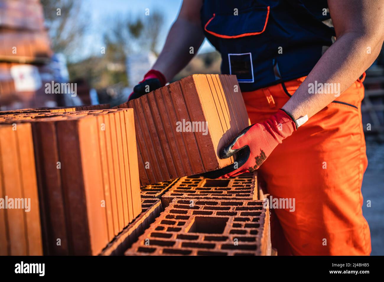 One man construction worker taking and carry or hold clay orange hollow blocks ar warehouse or construction site in sunny summer day copy space Stock Photo