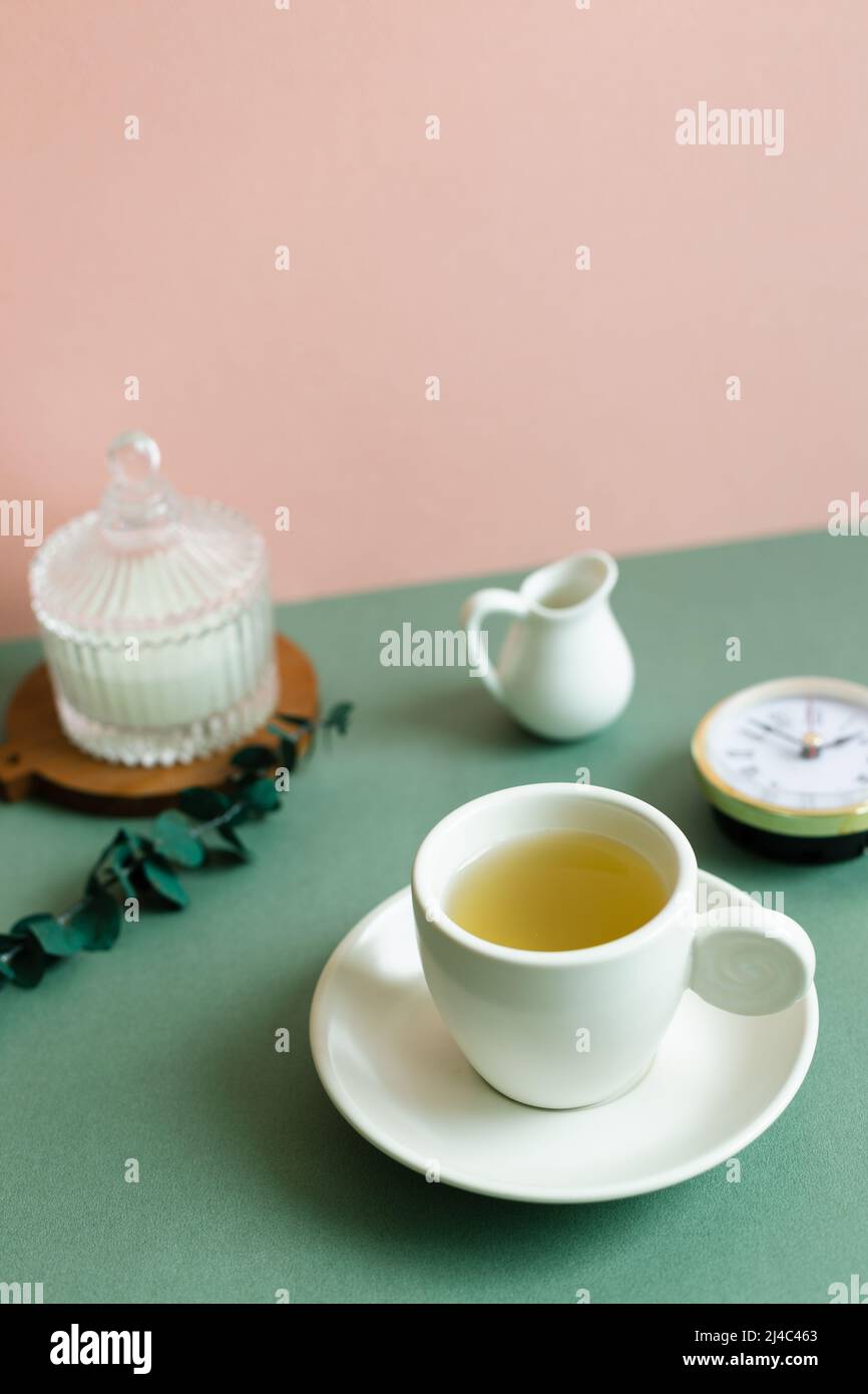Cup of tea, clock, candle, eucalyptus leaf on green table. pink wall ...