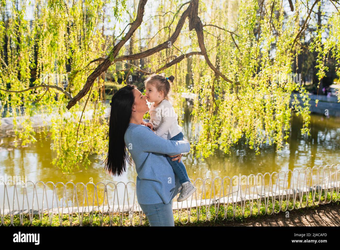 A casual clothed caucasian woman holds her raised daughter in her arms. A cute little girl with ponytails kisses her mother's nose. Enjoying spring day Stock Photo