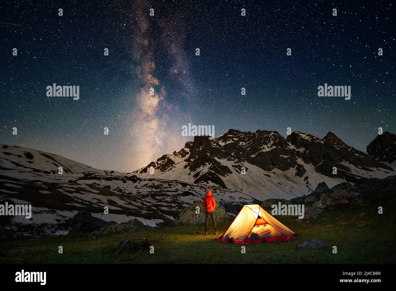 Hiking tourist standing near illuminated tent under milky way stars Stock Photo