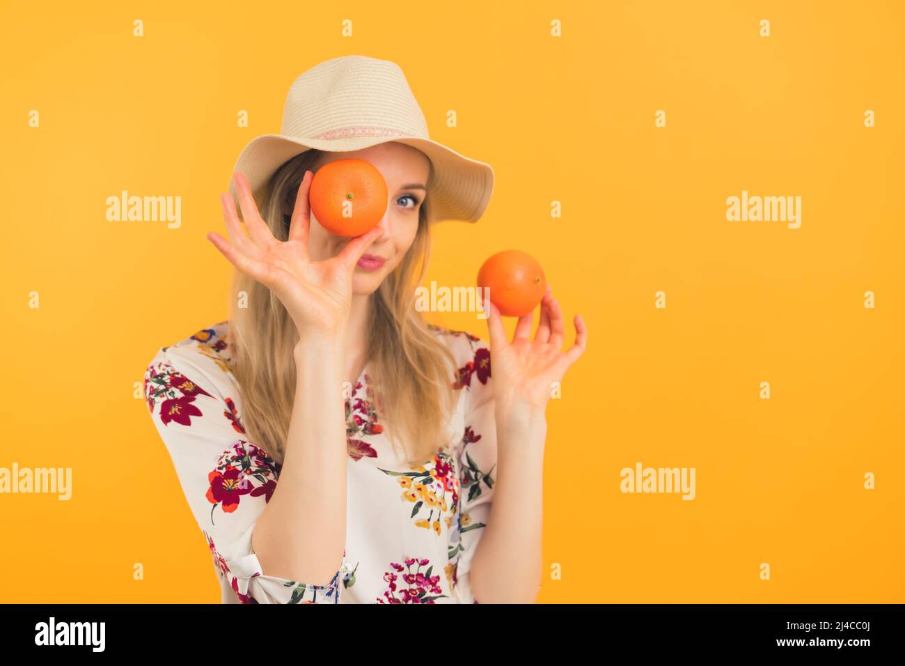 Youthful caucasian blonde girl in her 20s in a hat posing with two oranges or mandarins. Medium studio shot over orange background. High quality photo Stock Photo