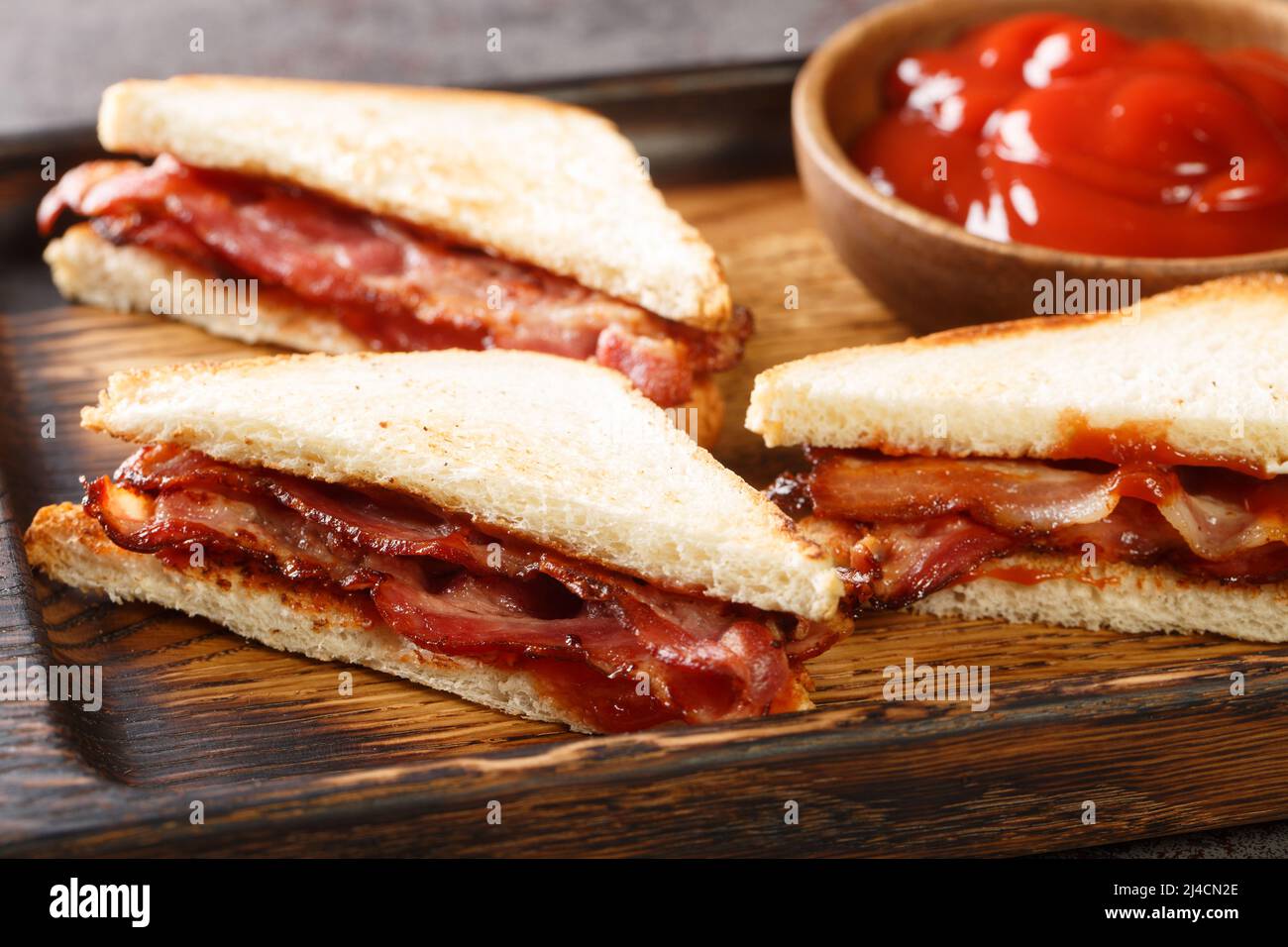 Bacon butty is a British sandwich consisting of crispy bacon, butter, and sauce closeup in the wooden tray on the table. Horizontal Stock Photo