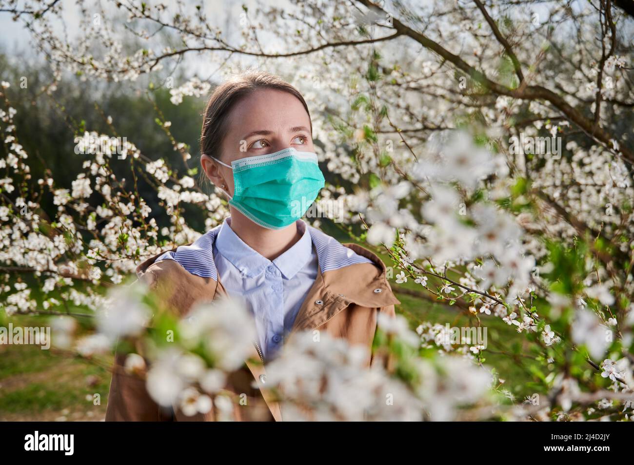 Woman allergic suffering from seasonal allergy at spring, posing in blossoming garden at springtime, wearing medical mask among blooming trees. Spring allergy concept Stock Photo