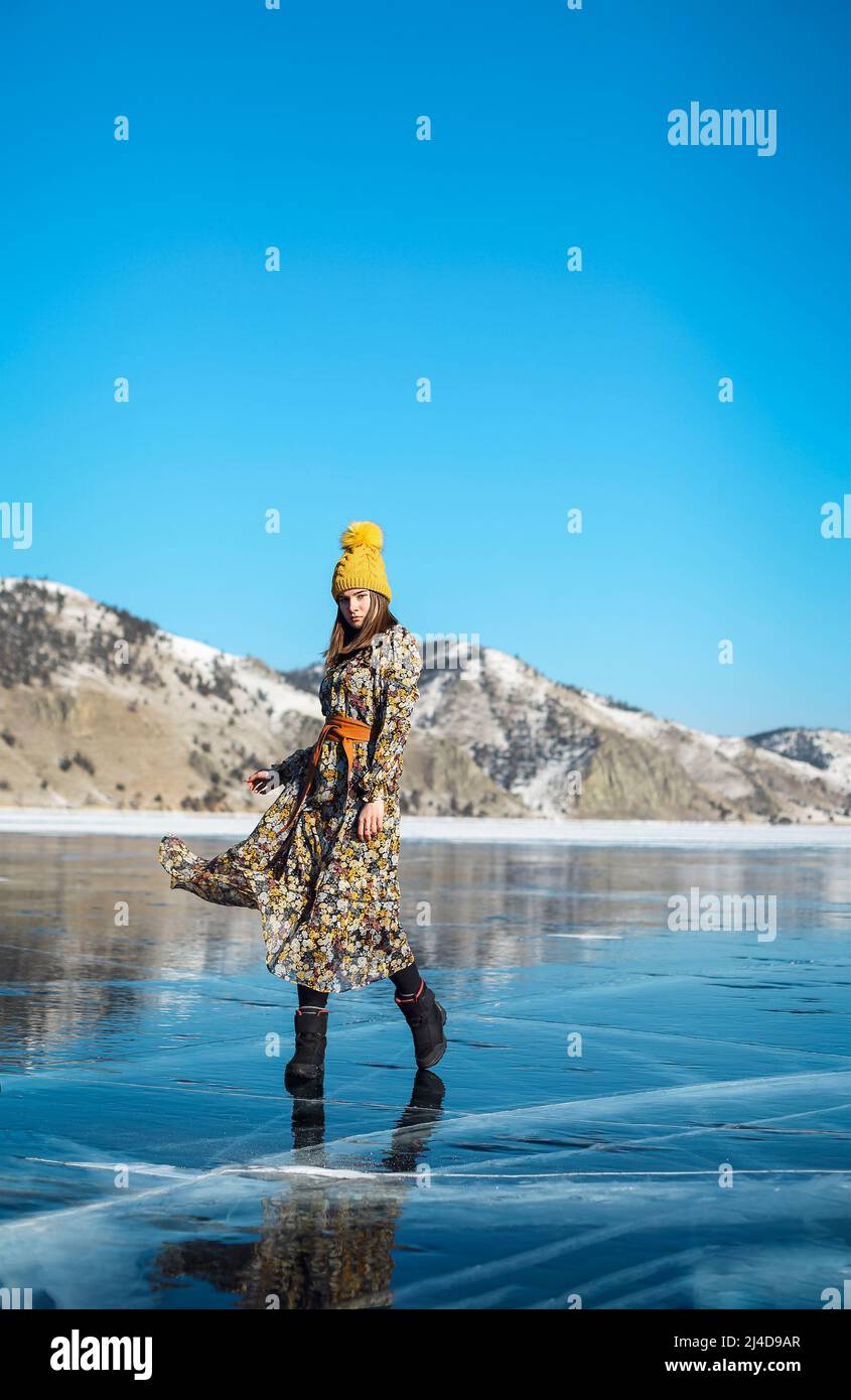 Fashionable image of a stunning woman posing on Baikal Lake ice wearing yellow hat. Boho dress flies in the wind. Beautiful ice landscapes of Lake Bai Stock Photo