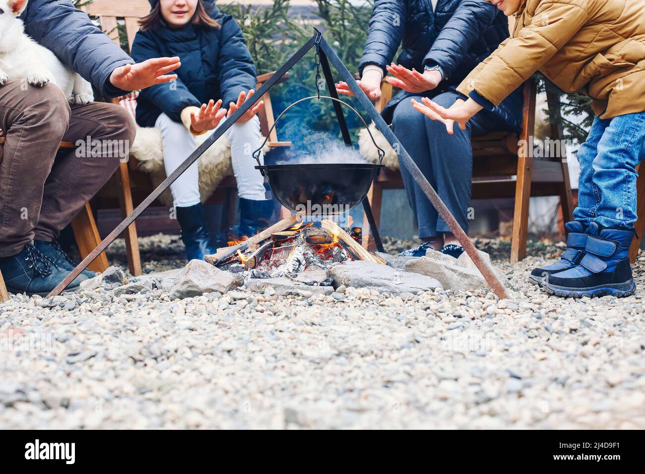 New Years gatherings by winter bonfire. Cozy portrait of happy family parents, two children and sweet dog cooking food in steaming pot over campfire w Stock Photo