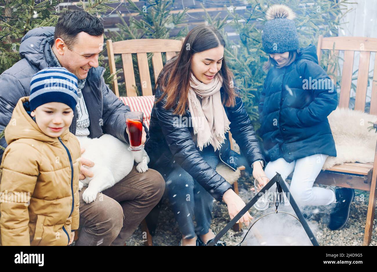 New Years gatherings by winter bonfire. Cozy portrait of happy family parents, two children and sweet dog cooking food in steaming pot over campfire w Stock Photo