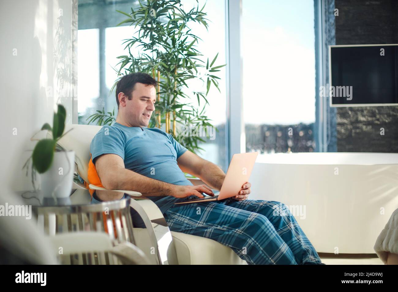 Handsome young smiling man in casual pajamas outfit sitting in armchair in morning working on laptop from his bedroom, freelancer at home Stock Photo