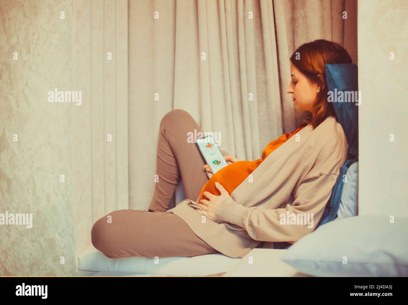 Pregnant woman holding smartphone using pregnancy tracker app on mobile phone while sitting indoors, expectant mother tracking baby development in mob Stock Photo