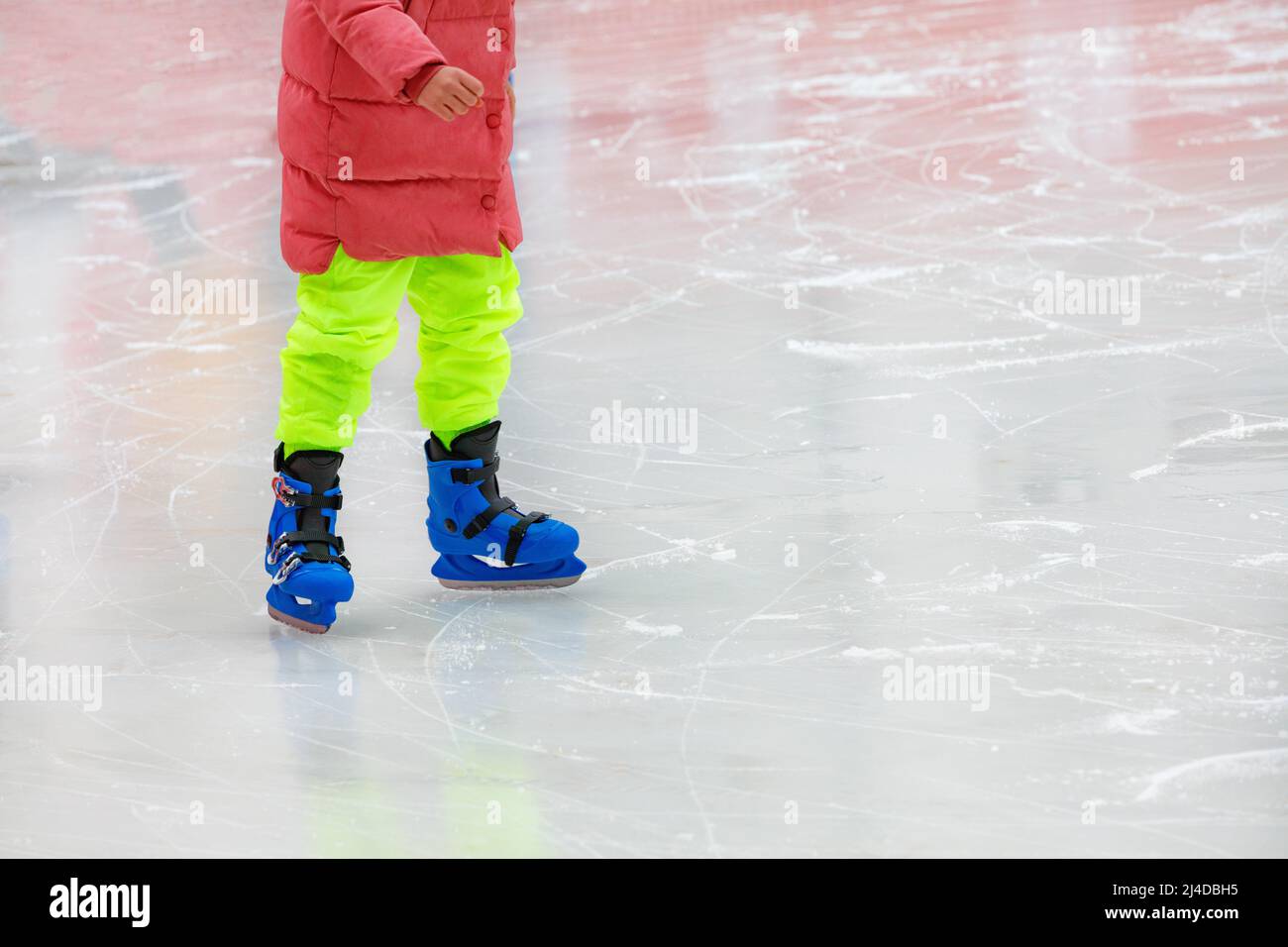 A teenager in bright green sweatpants, a red down jacket and blue skates learns to skate. Copy space, selective focus, close-up. Stock Photo